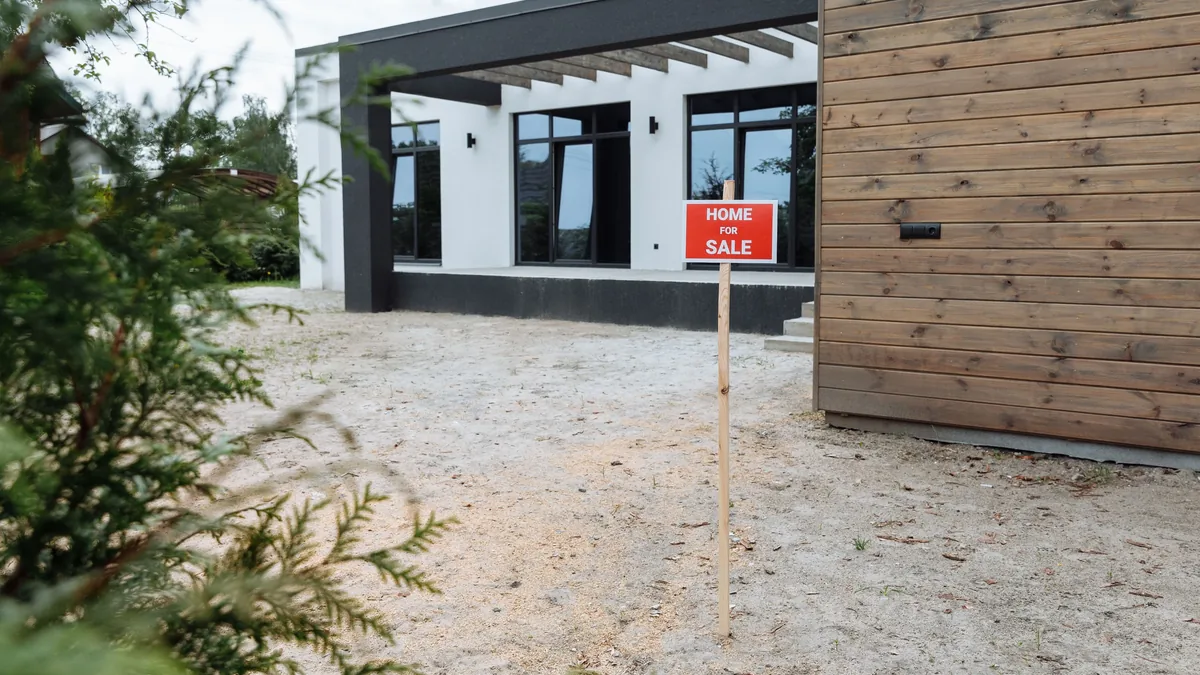 A modern house with a red "HOME SALE" sign in front of it, situated on a sandy lot with a wooden wall and a tree in the foreground.